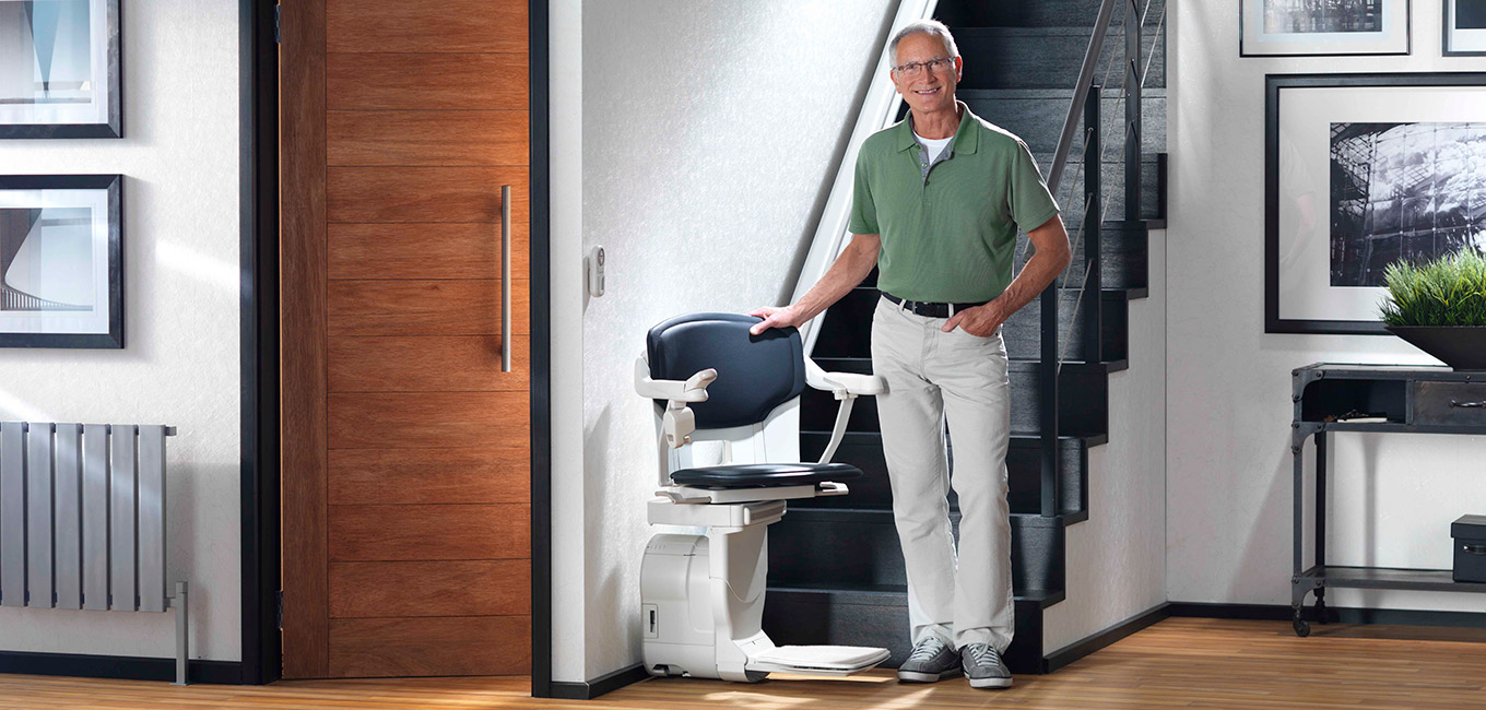 Umfassendes Sortiment an Treppenlifte, Rollstuhllifte und Homelifte, optimale Lösungen für ein barrierefreies Zuhause. Ein älterer Herr steht neben dem Stuhltreppenlift an der Treppe